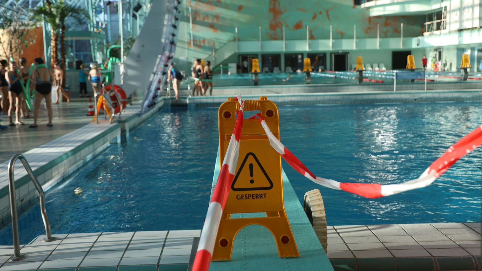 Ein Schild mit der Aufschrift „geperrt“ steht auf einem Sprungbrett. Im Hintergrund sieht man ein Schwimmbecken, um das Menschen laufen.