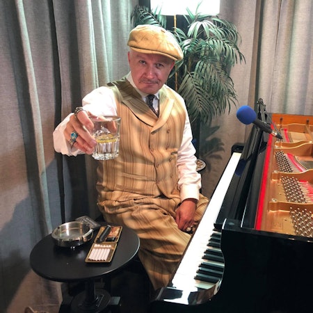 Christian Christl sitzt am Piano und hält ein Glas in die Kamera.
