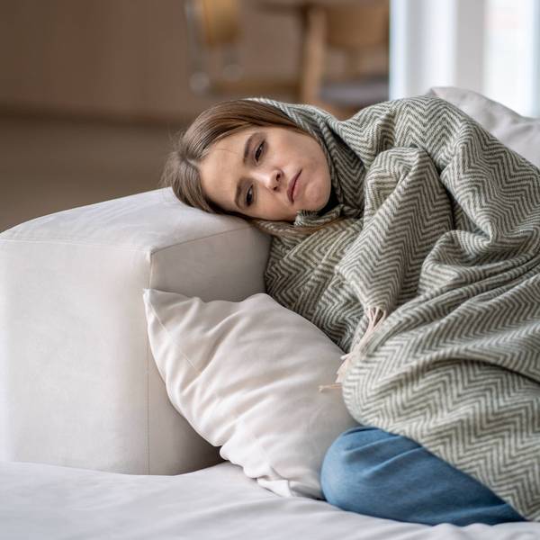 Traurig guckende Frau liegt umhüllt von einer Decke auf dem Sofa.