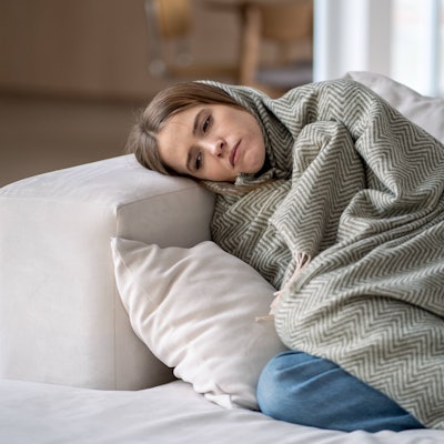 Traurig guckende Frau liegt umhüllt von einer Decke auf dem Sofa.