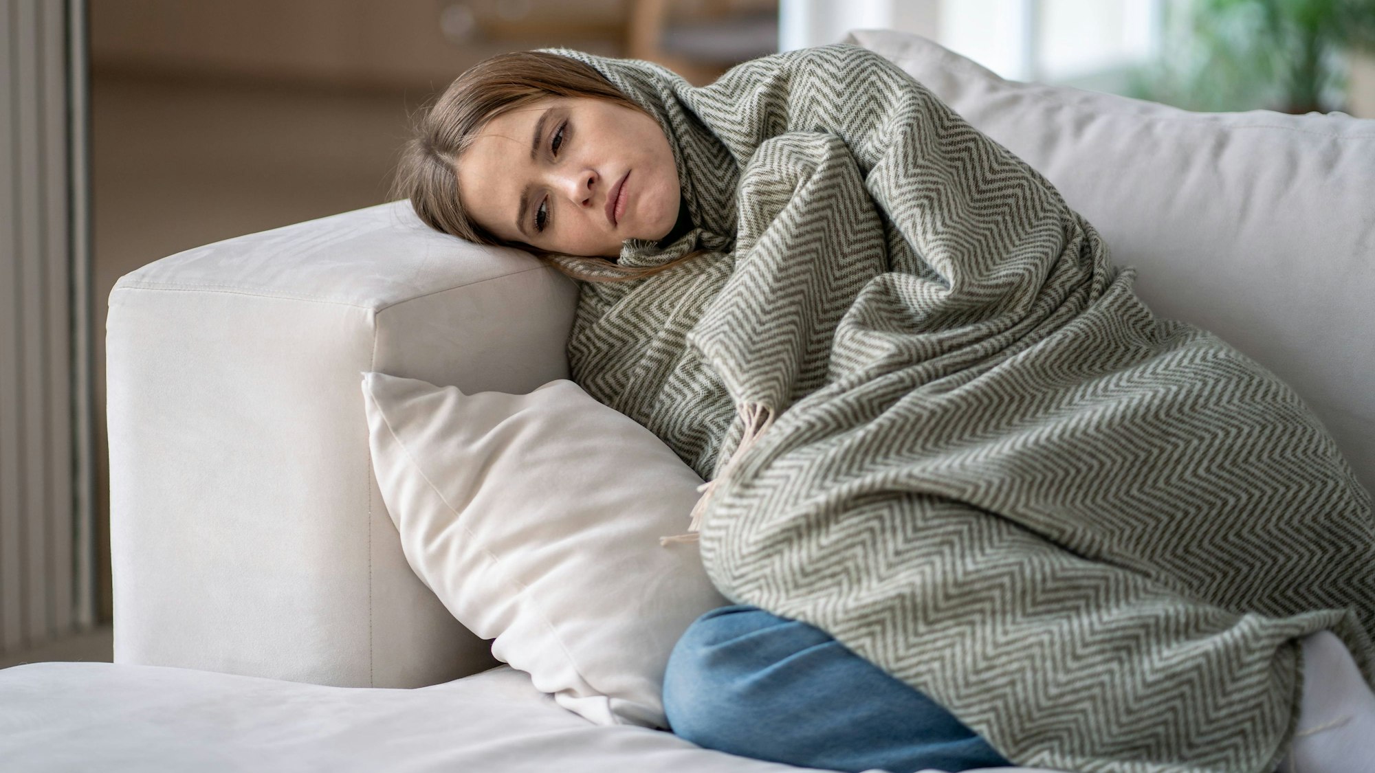 Traurig guckende Frau liegt umhüllt von einer Decke auf dem Sofa.