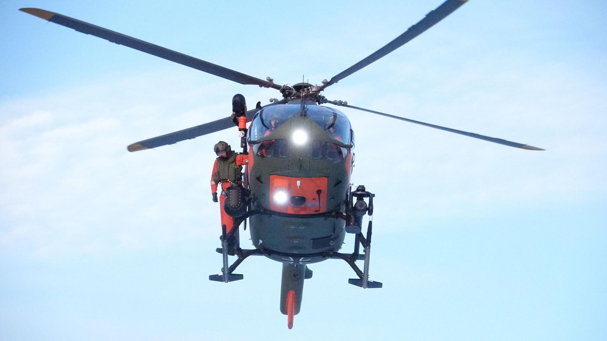 Zu sehen ist ein Hubschrauber der Bundeswehr im Flug, ein Besatzungsmitglied steht außen auf einer Kufe.