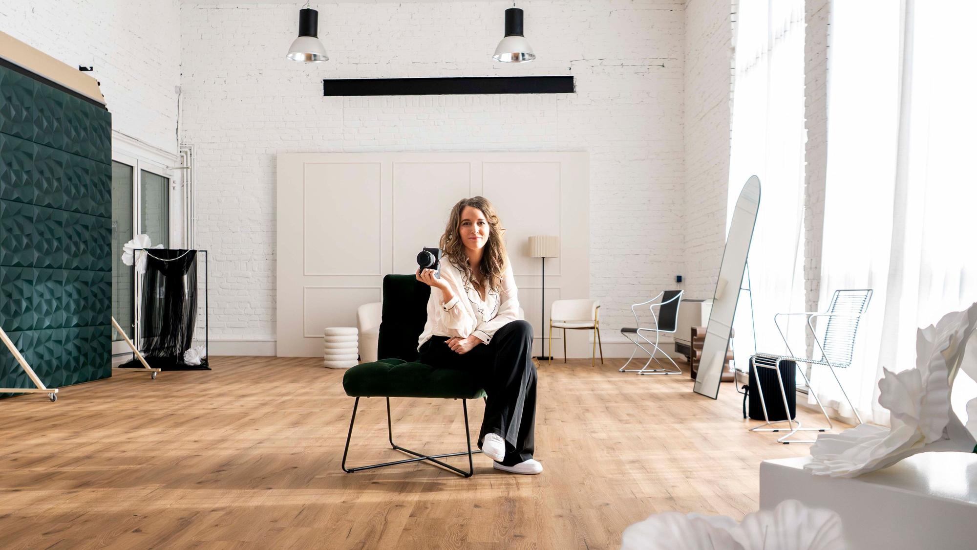 Sarah Pfeiffer-Schröder sitzt in ihrem Studio auf einem Stuhl, in der Hand hält sie eine Kamera.