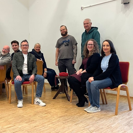 Neun Menschen sitzen auf Stühlen oder stehen in einem Pfarrzentrum.