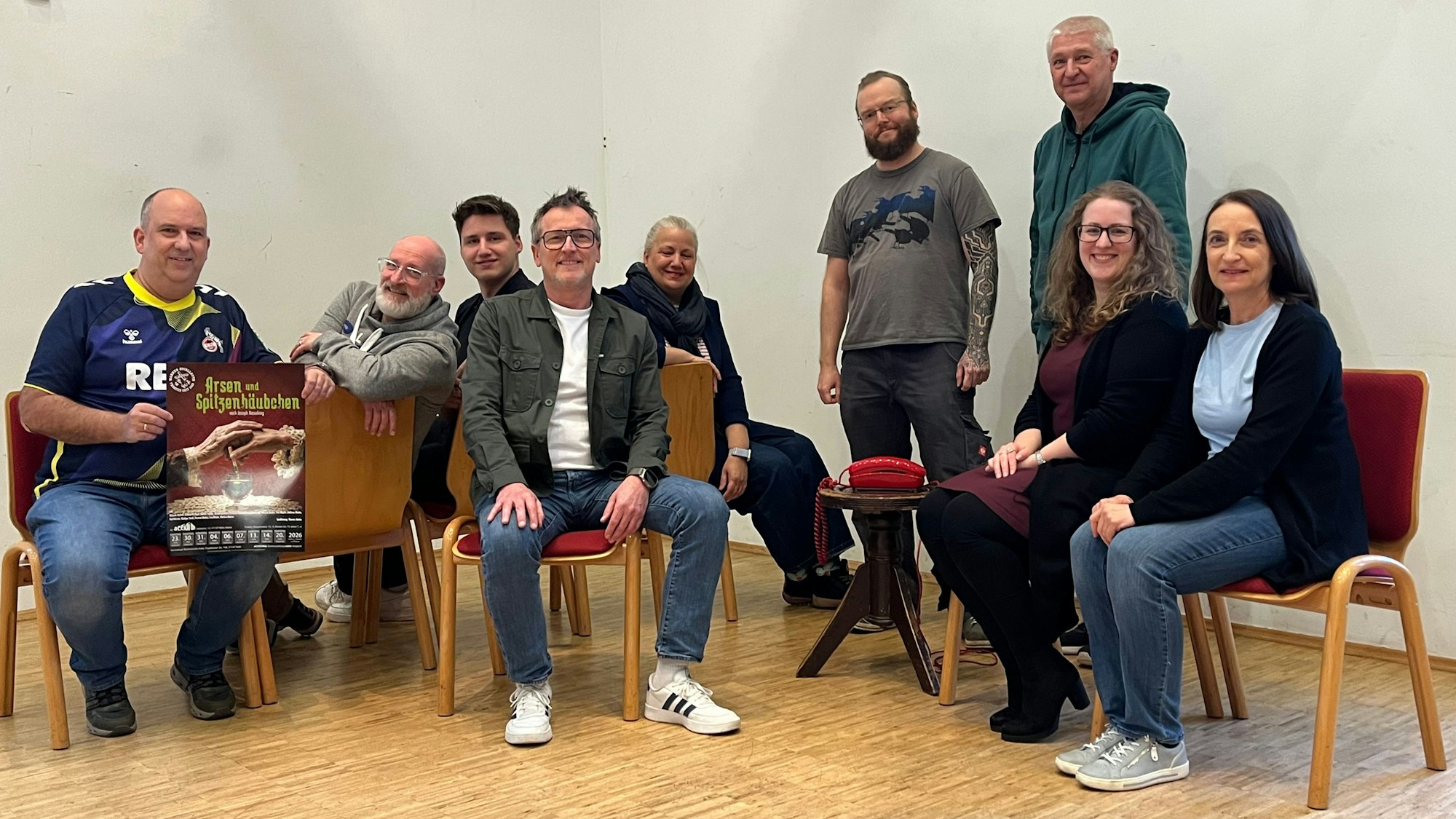 Neun Menschen sitzen auf Stühlen oder stehen in einem Pfarrzentrum.