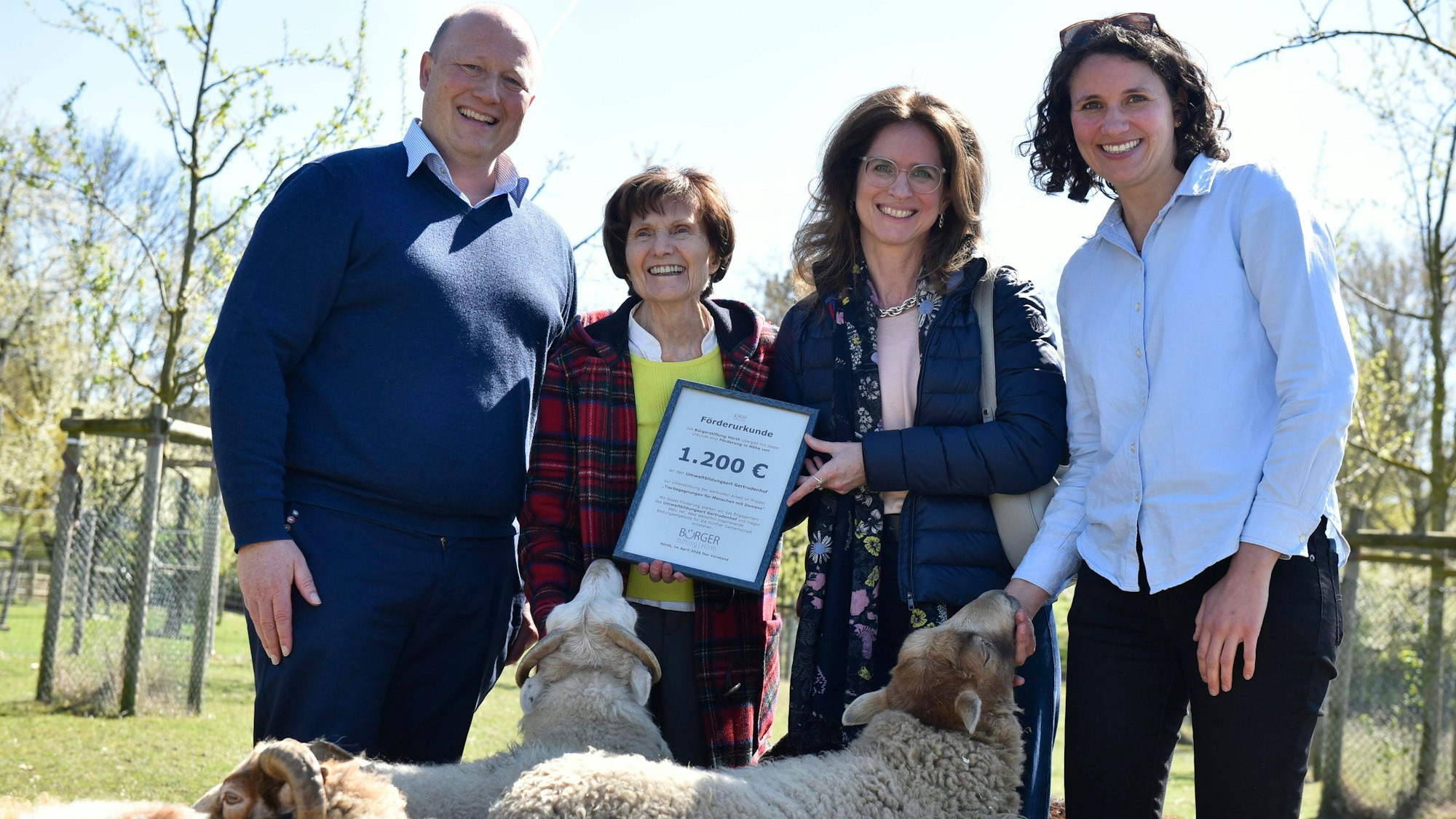 Drei Frauen und ein Mann lächeln in die Kamera, eine Schafherde umringt sie.