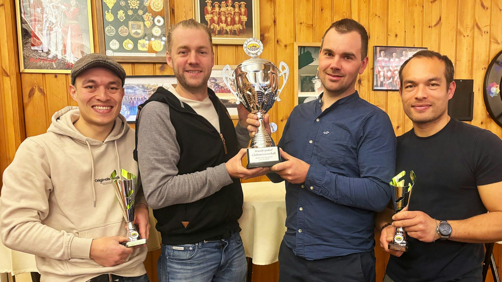 Vier Männer stehen nebeneinander vor einer holzvertäfelten Wand. Drei von ihnen halten Pokale in den Händen.