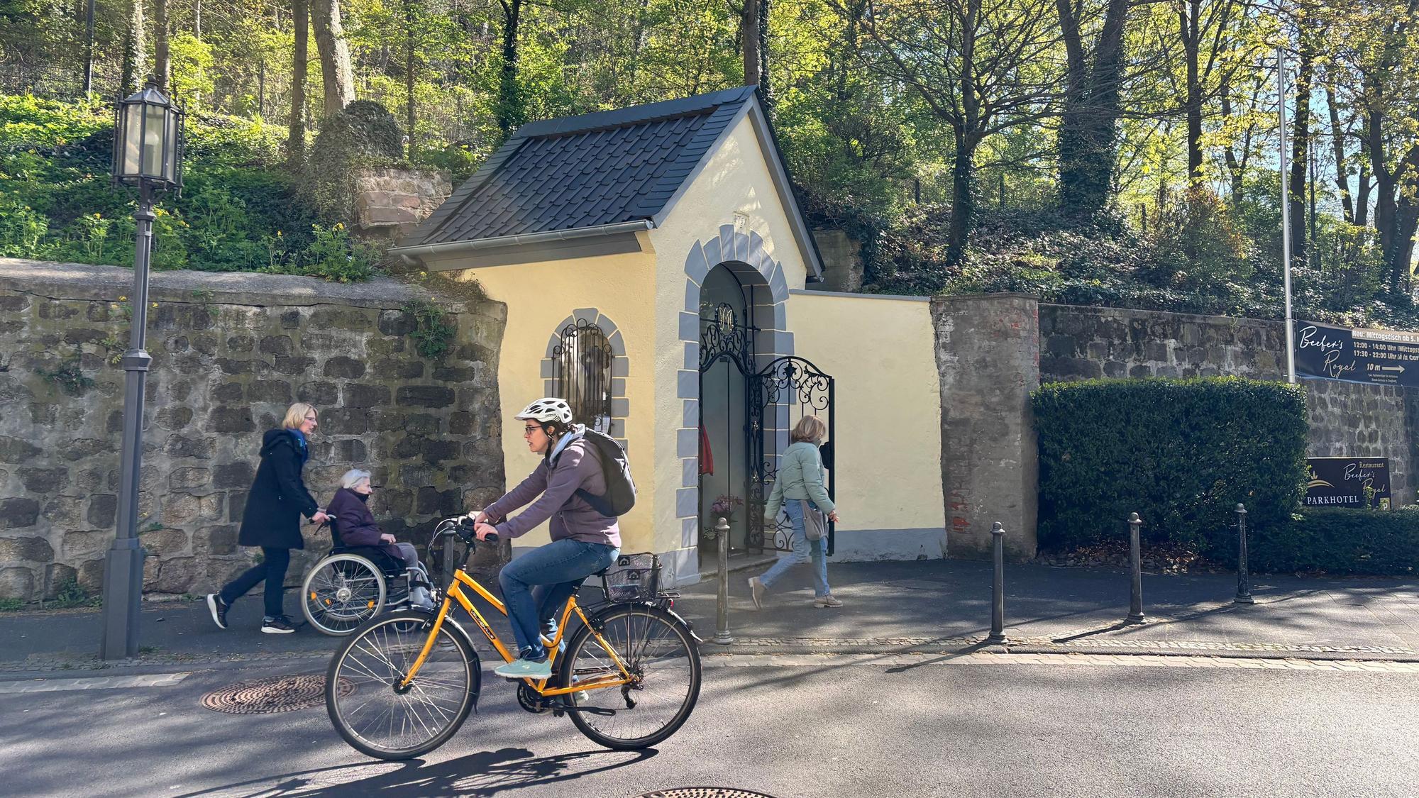 Vor einer kleinen Kapelle fährt eine Radfahrerin auf der Straße. Im Hintergrund sind weitere Menschen unterwegs.