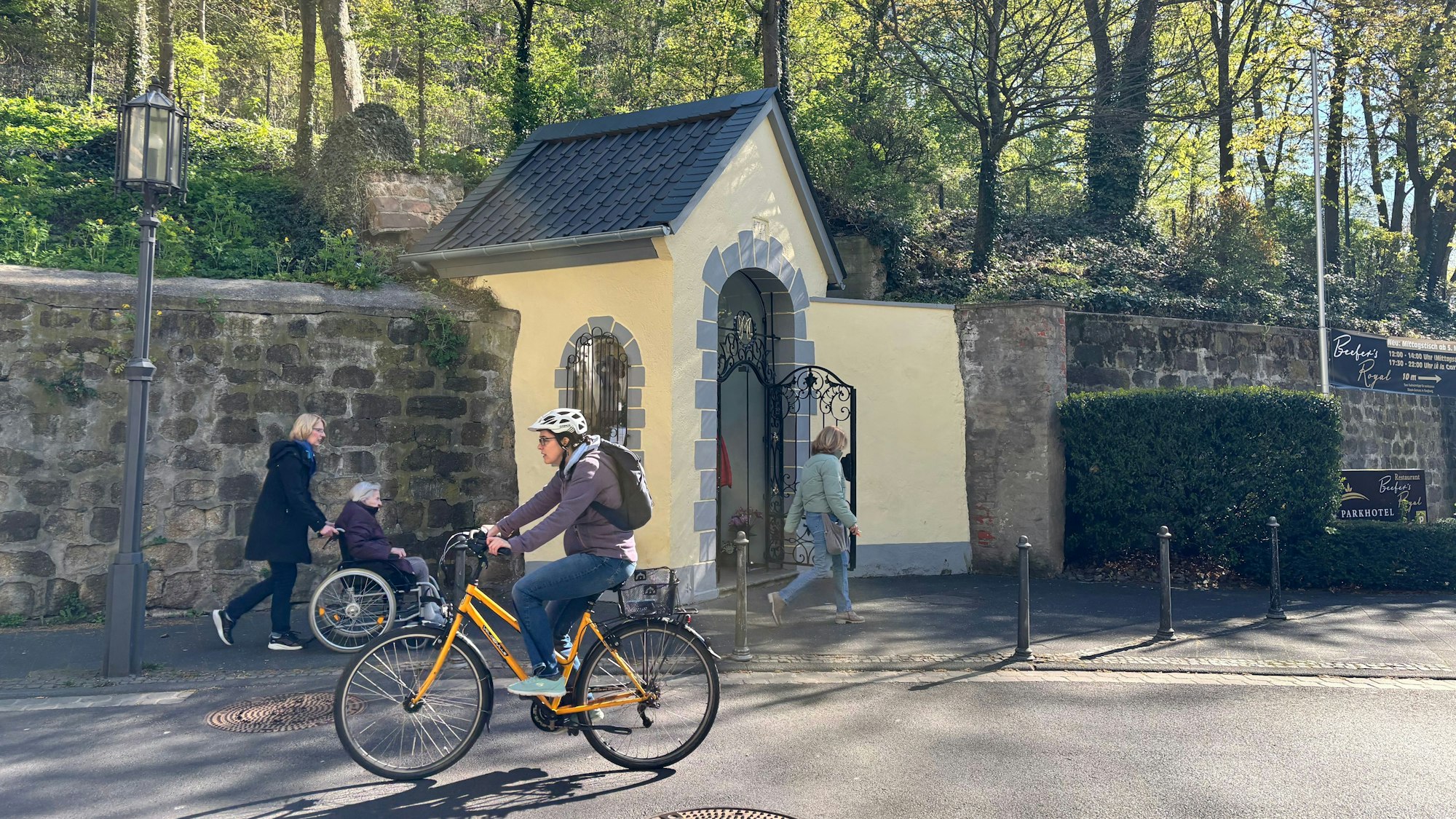 Vor einer kleinen Kapelle fährt eine Radfahrerin auf der Straße. Im Hintergrund sind weitere Menschen unterwegs.