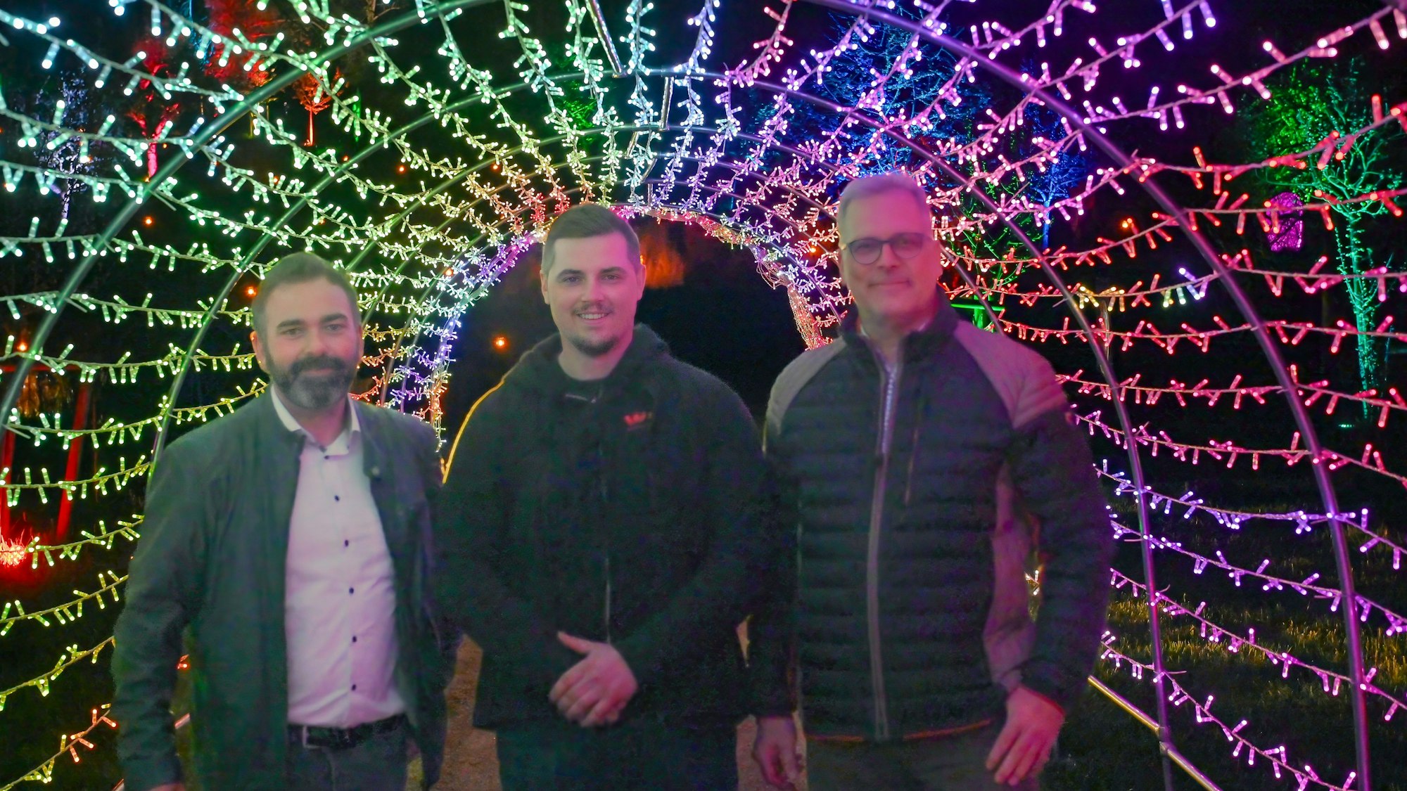 Die oben Genannten stehen in einem Tunnel aus bunten Lichtern.