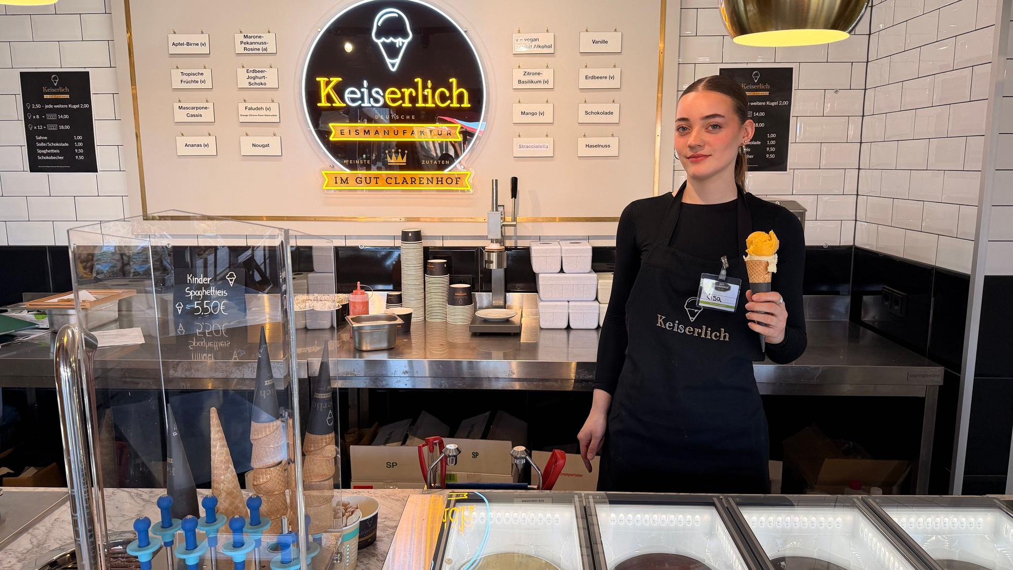 Eine junge Frau hält ein Eis in einer Waffel in der Hand, vor ihr die Verkaufstheke.