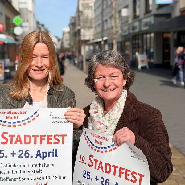 Die beiden Frauen stehen in der belebten Fußgängerzone, in den Händen halten sie jeweils ein Plakat für das Stadtfest.