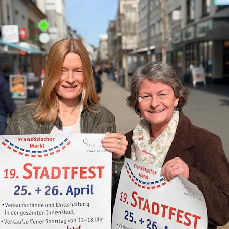 Die beiden Frauen stehen in der belebten Fußgängerzone, in den Händen halten sie jeweils ein Plakat für das Stadtfest.