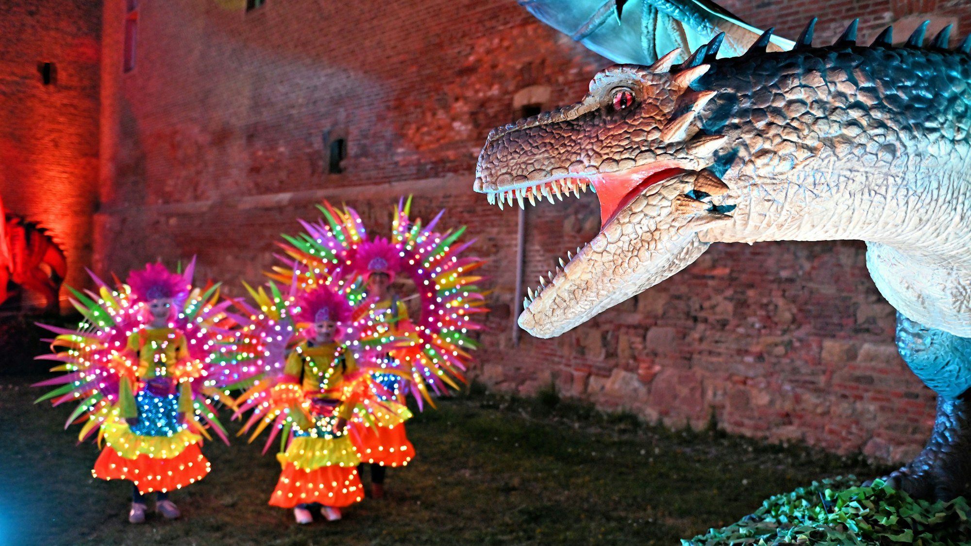 Vom rechten Bildrand droht ein Drache, links tanzen Menschen in bunten Lichterkostümen.