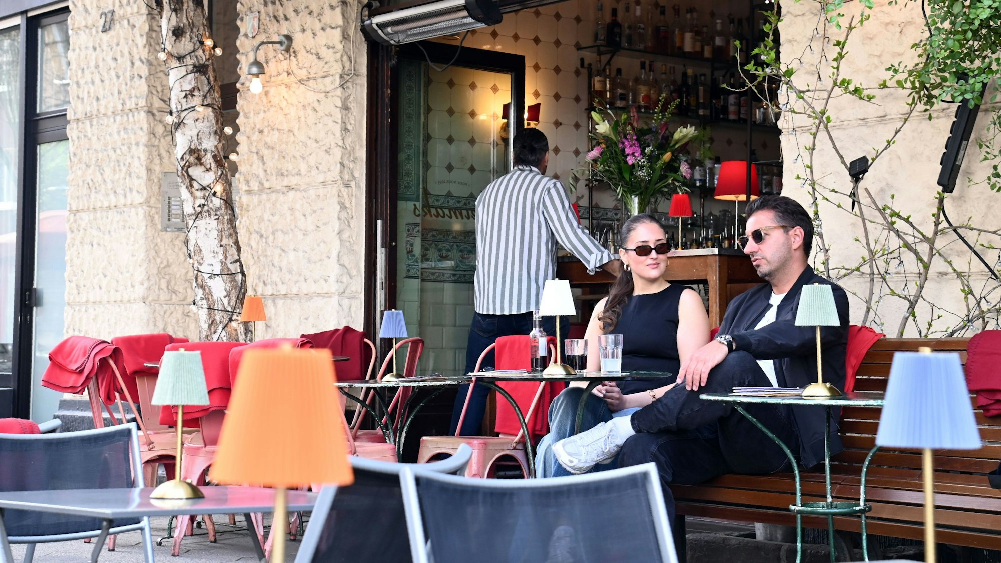 Der Außenbereich des Keimaks: Zwei Gäste sitzen auf einer Bank. Rote Decken sind ausgelegt. Auf den Tischen stehen bunte Tischleuchten.