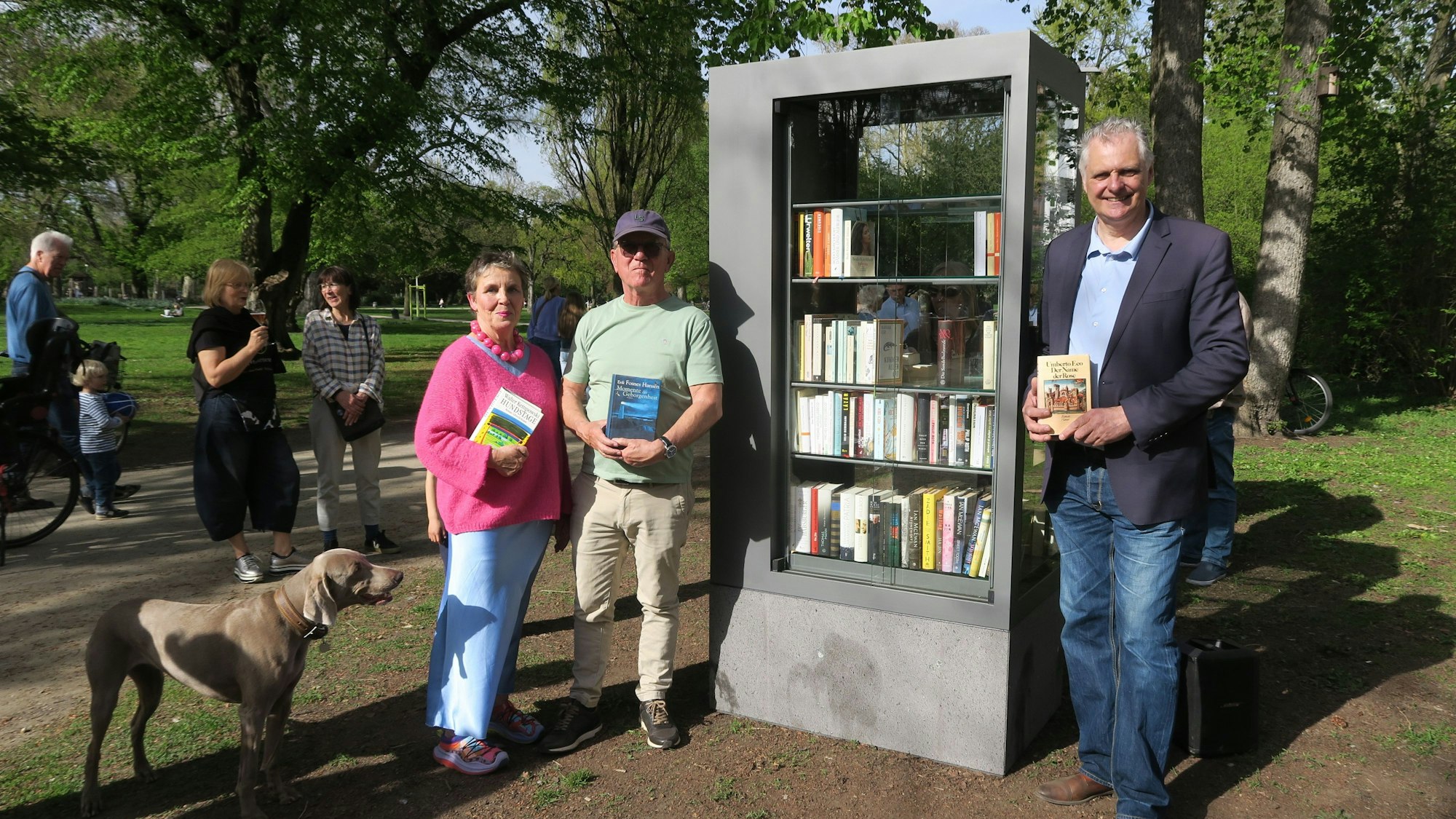 Drei Menschen mit Büchern in den Händen stehen neben einem neuen Bücherschrank im Kölner Stadtgarten. Daneben steht ein Hund und im Hintergrund sind weitere Menschen zu sehen.