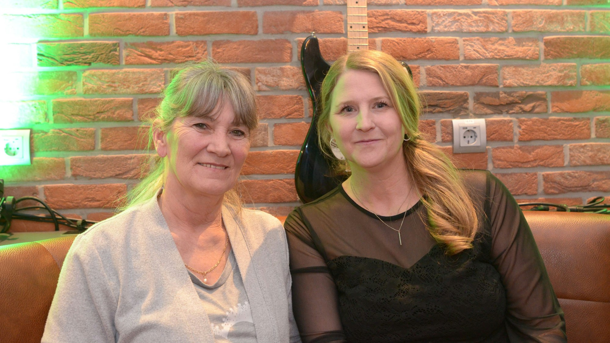 Zwei Frauen sitzen nebeneinander auf einer Bank in einem Café. An der Wand steht eine Gitarre als Dekoration.