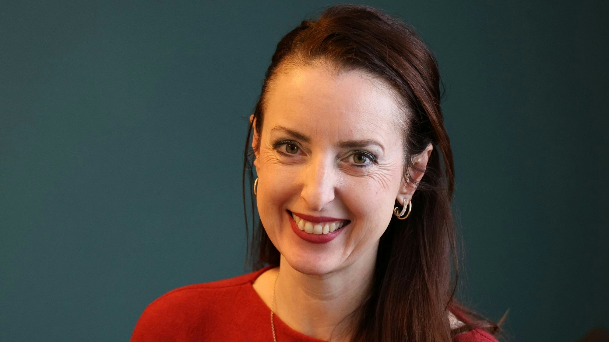 Eine Frau sitzt in einem roten Pullover und mit einem Buch in der Hand vor einer dunklen Wand.