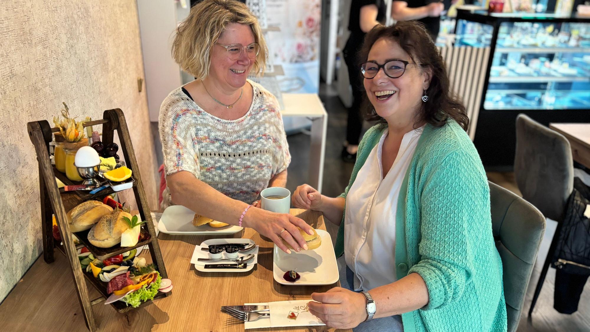 Zwei Frauen sitzen lachend an einem Café-Tisch, auf dem ein Frühstück aufgebaut ist.