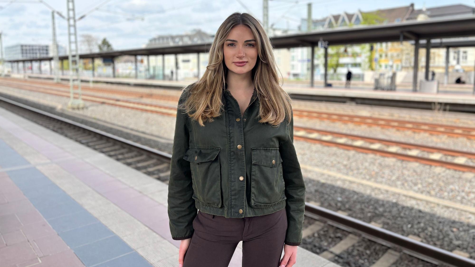 Janina Stifter steht auf einem Bahnsteig. Im Hintergrund verlaufen unscharf gestellten Schienen und stehen Häuser.