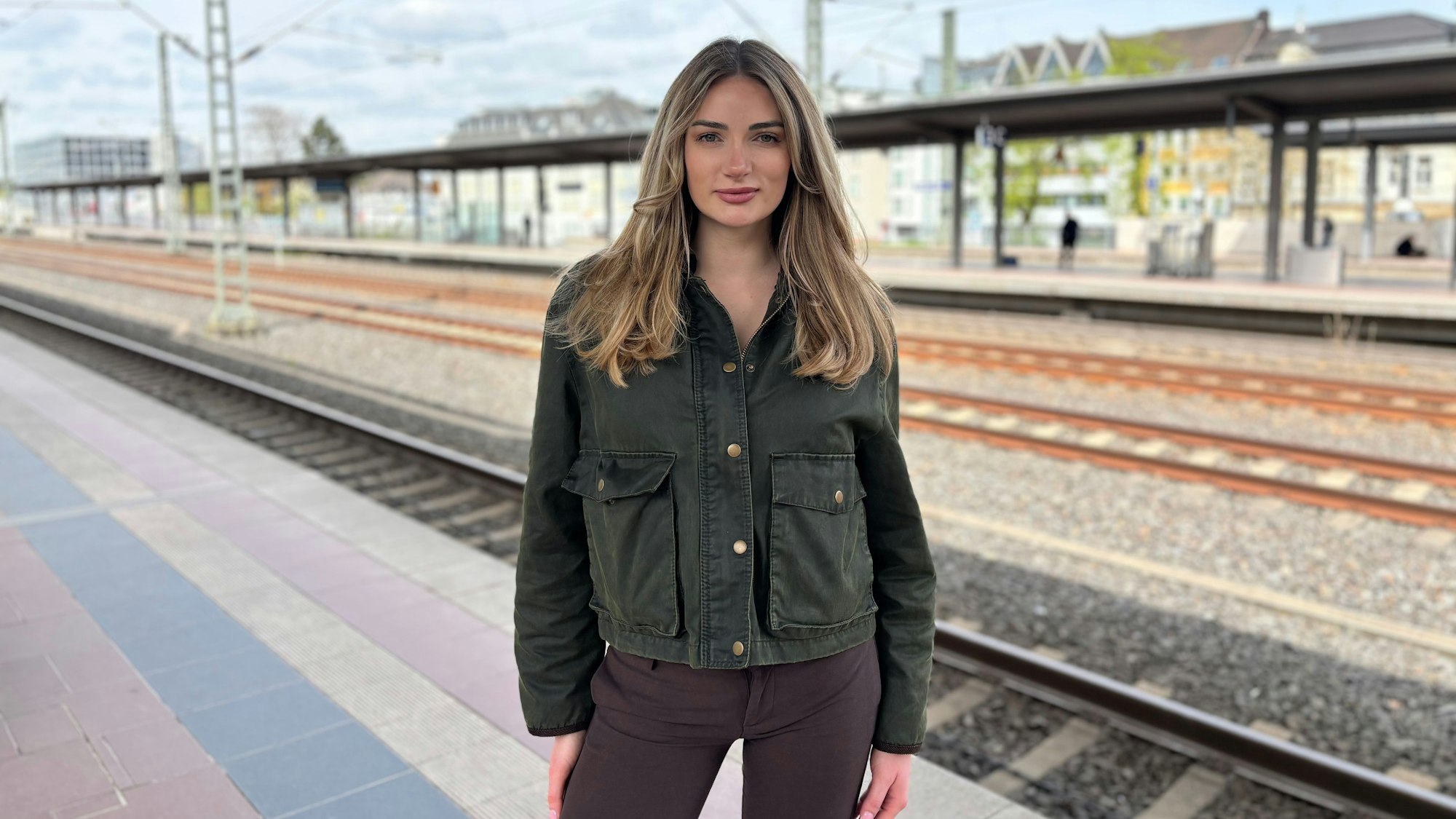 Janina Stifter steht auf einem Bahnsteig. Im Hintergrund verlaufen unscharf gestellten Schienen und stehen Häuser.