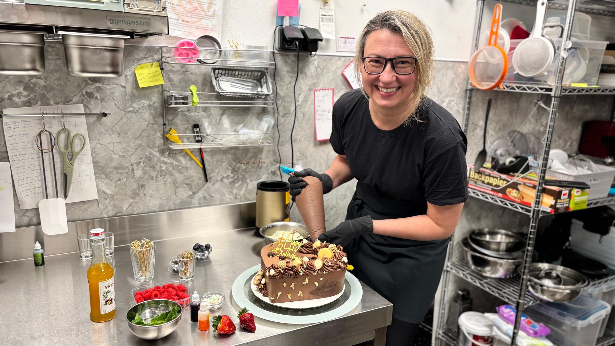 Eine Frau mit blonden Harren steht in schwarz gekleidet in einer Küche und verziert eine Schokoladen-Torte.