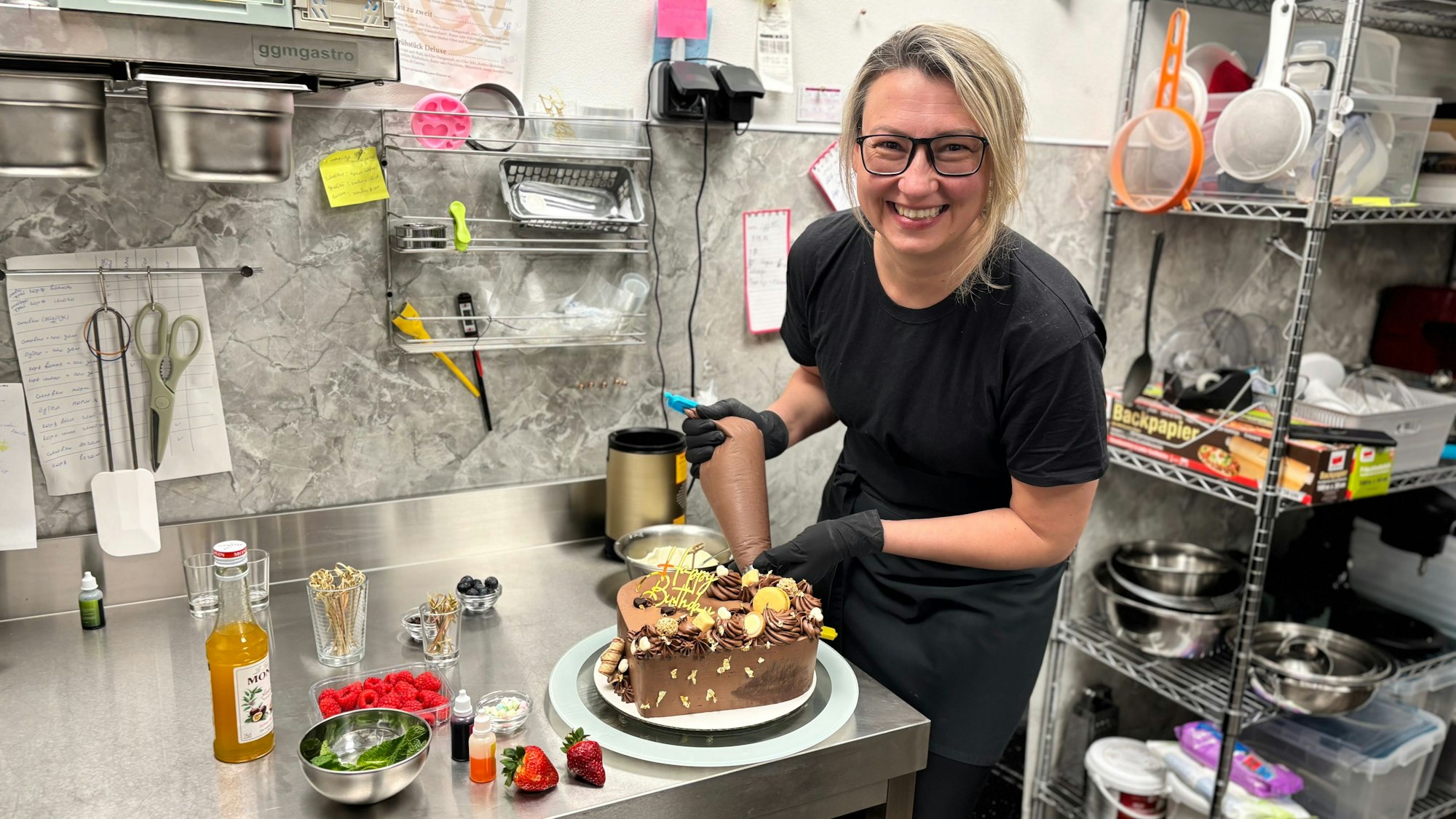 Eine Frau mit blonden Harren steht in schwarz gekleidet in einer Küche und verziert eine Schokoladen-Torte.
