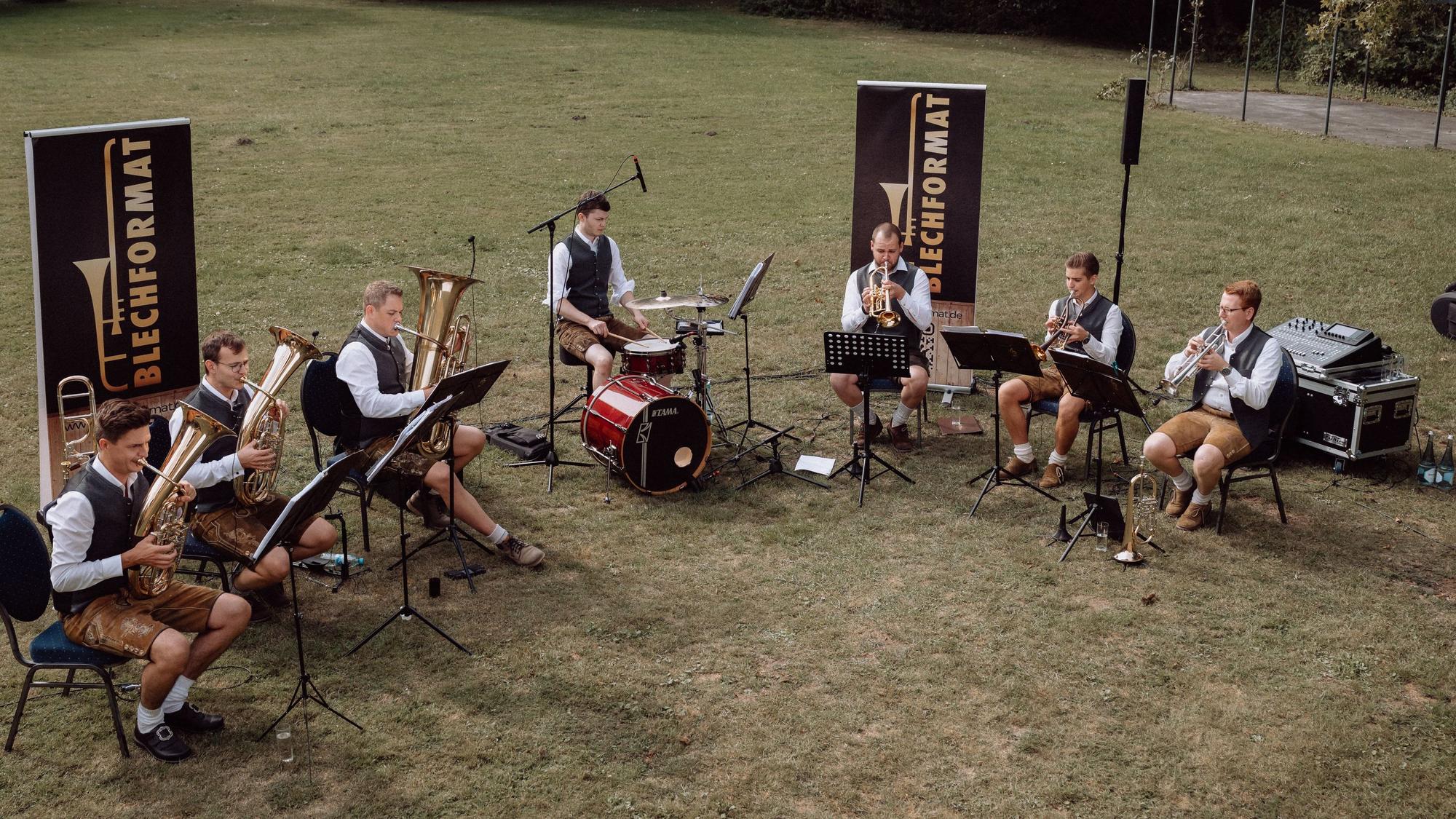 Die Musiker der Gruppe Blechformat sitzen im Halbkreis auf Stühlen auf einer Wiese und machen Musik.