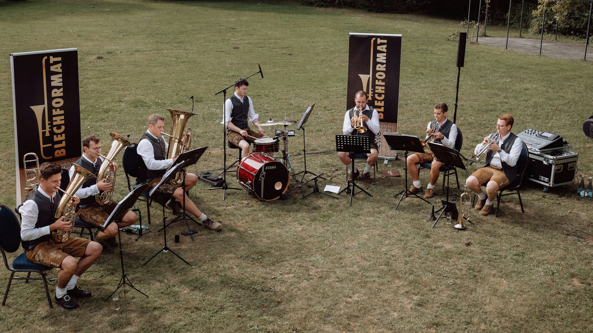 Die Musiker der Gruppe Blechformat sitzen im Halbkreis auf Stühlen auf einer Wiese und machen Musik.