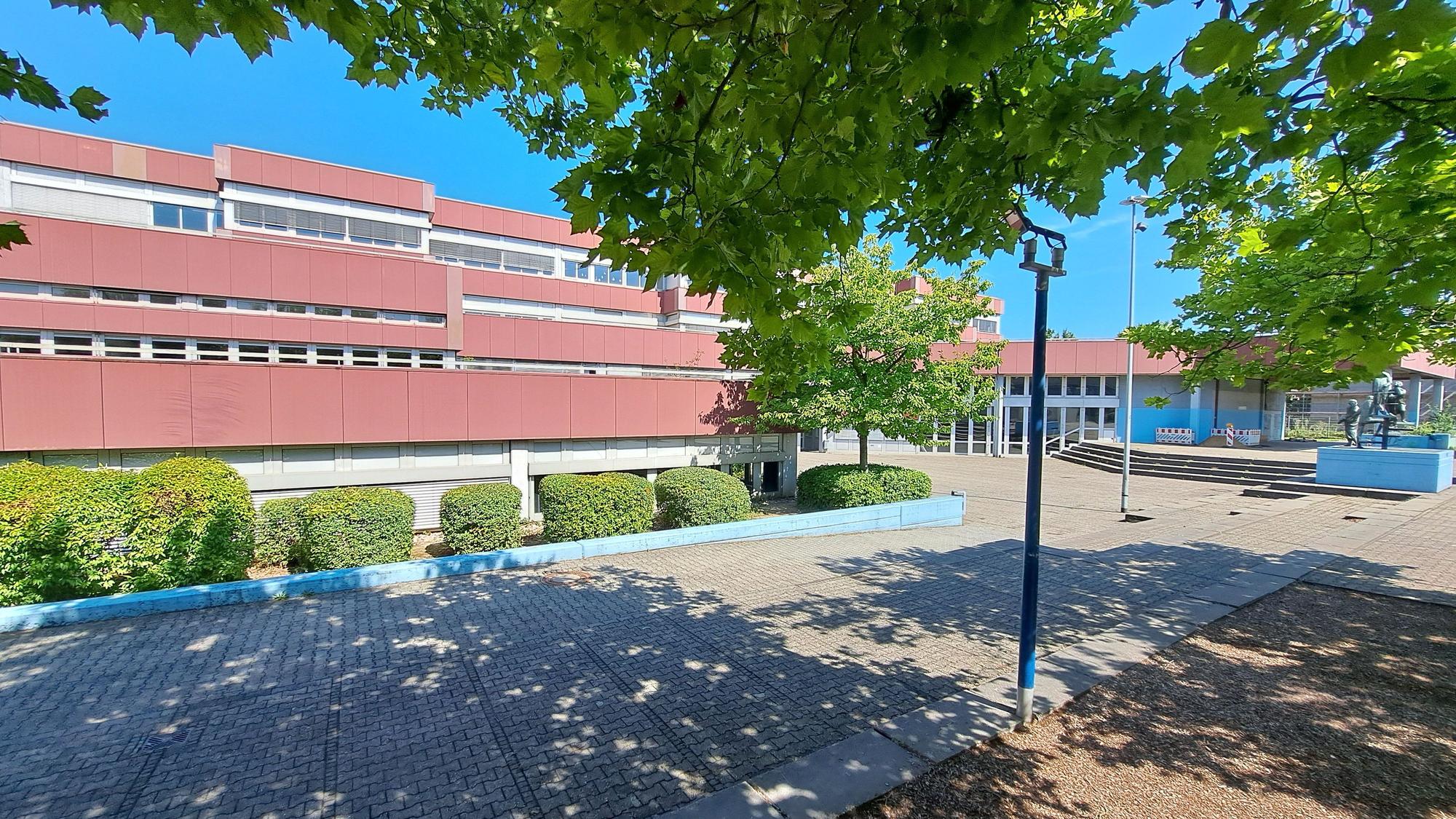 Die Aufnahme zeigt das Kopernikus-Gymnasium im Niederkasseler Schulzentrum Nord. Im Vordergrund werfen Bäume einen Schatten auf Pflastersteine.