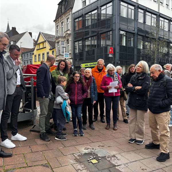 Menschen stehen in einem Halbkreis um den Stolperstein und blicken zu Boden.