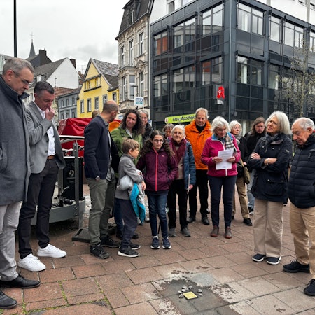 Menschen stehen in einem Halbkreis um den Stolperstein und blicken zu Boden.
