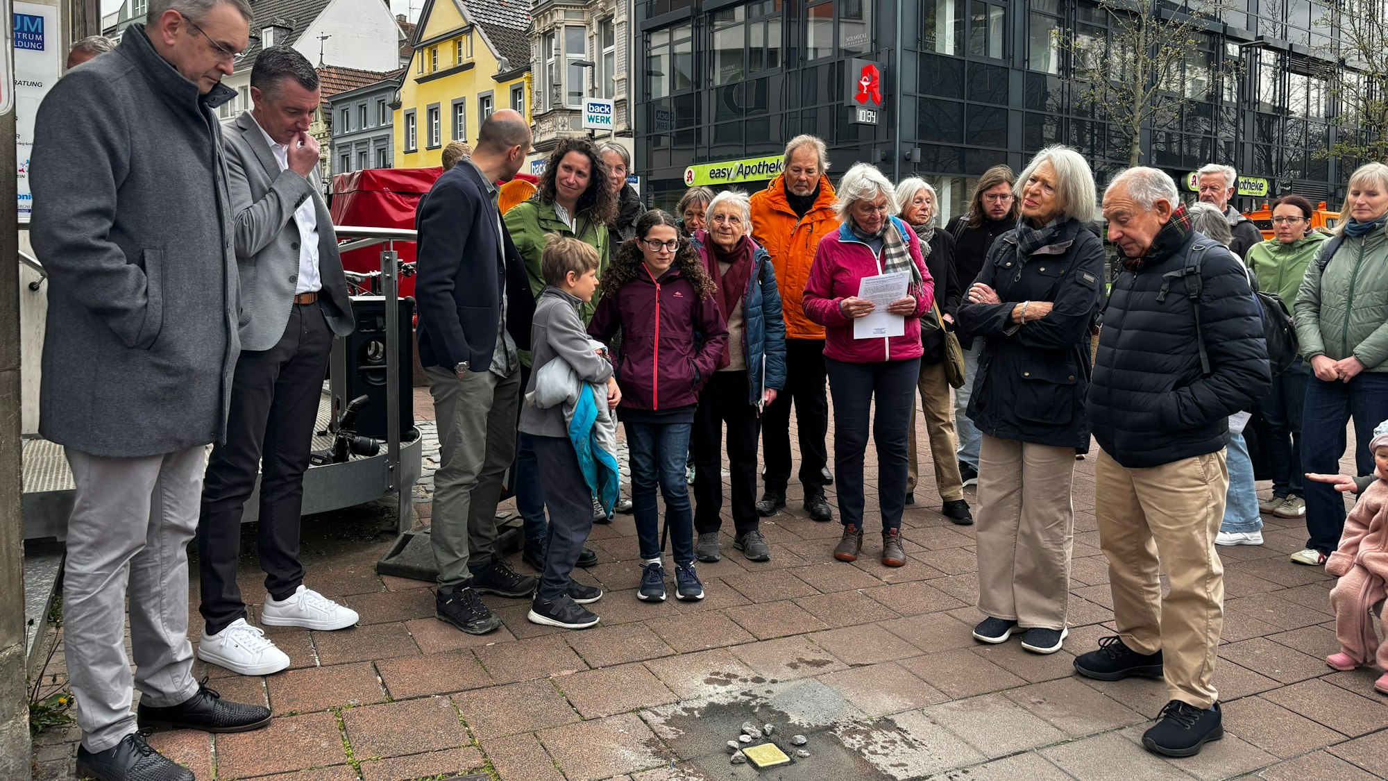 Menschen stehen in einem Halbkreis um den Stolperstein und blicken zu Boden.