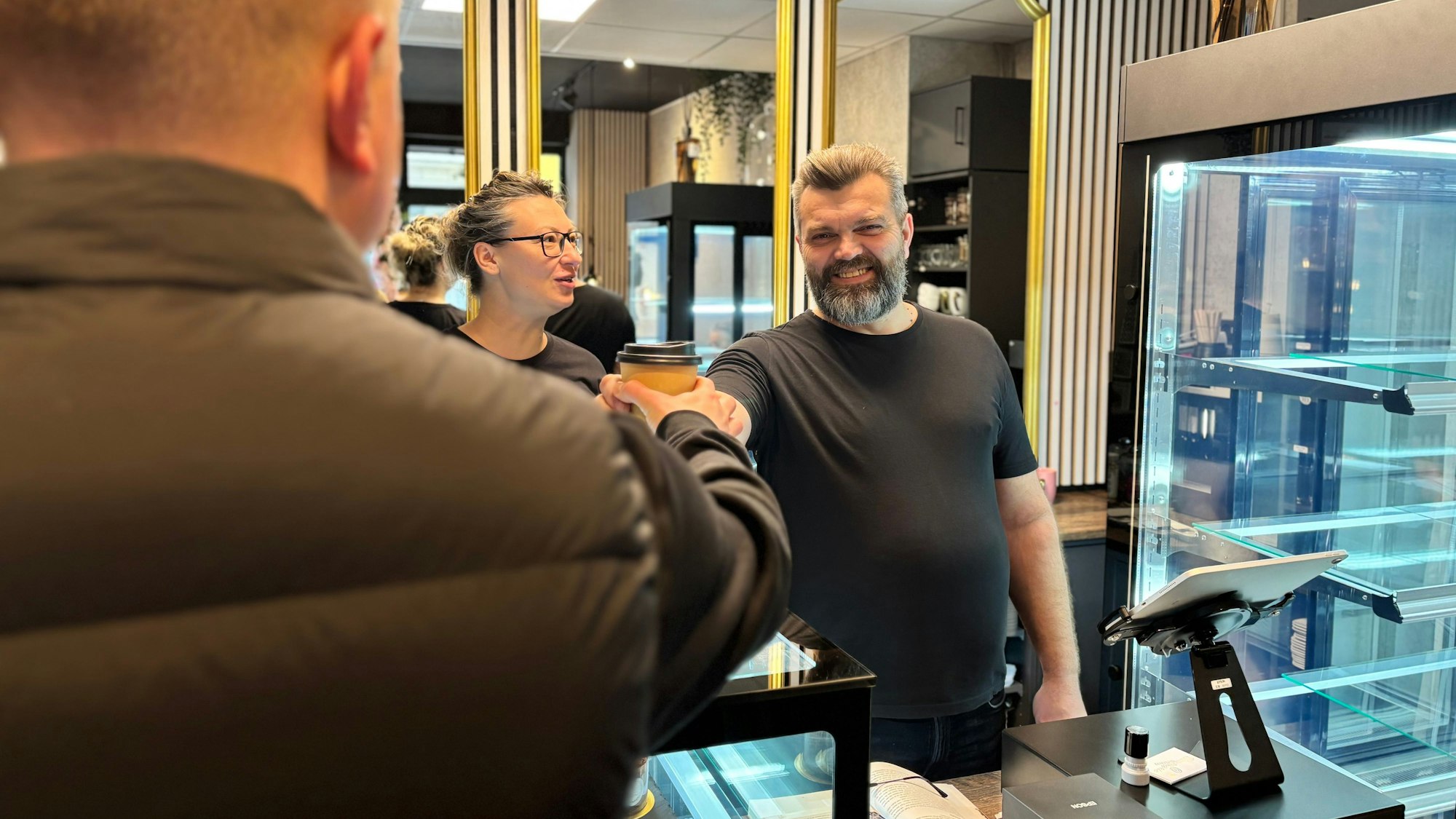 Ein Mann mit Bart und in schwarzem T-Shirt reicht einem Kunden einen Kaffee.