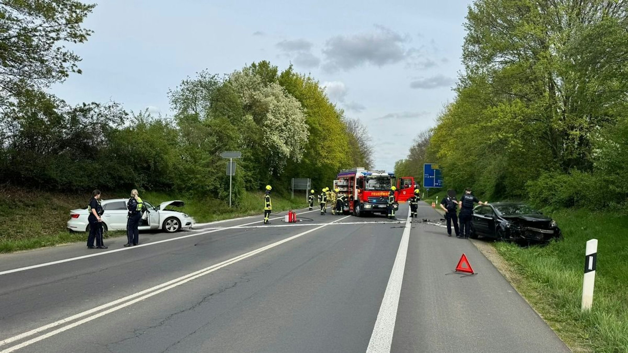 Ein bei einem Unfall beschädigtes Auto steht auf einer Straße, Feuerwehrleute sind im Hintergrund zu sehen.