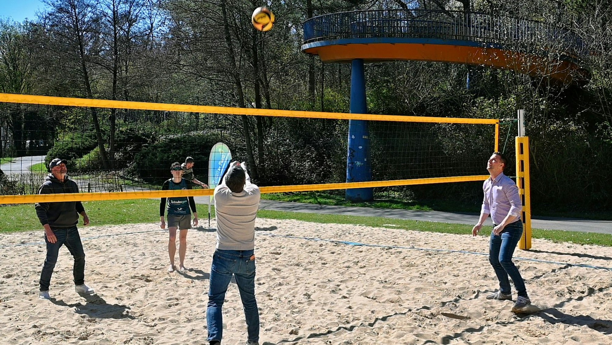 Auf einem Spielfeld mit Sandboden sieht man vier Menschen Beachvolleyball spielen.