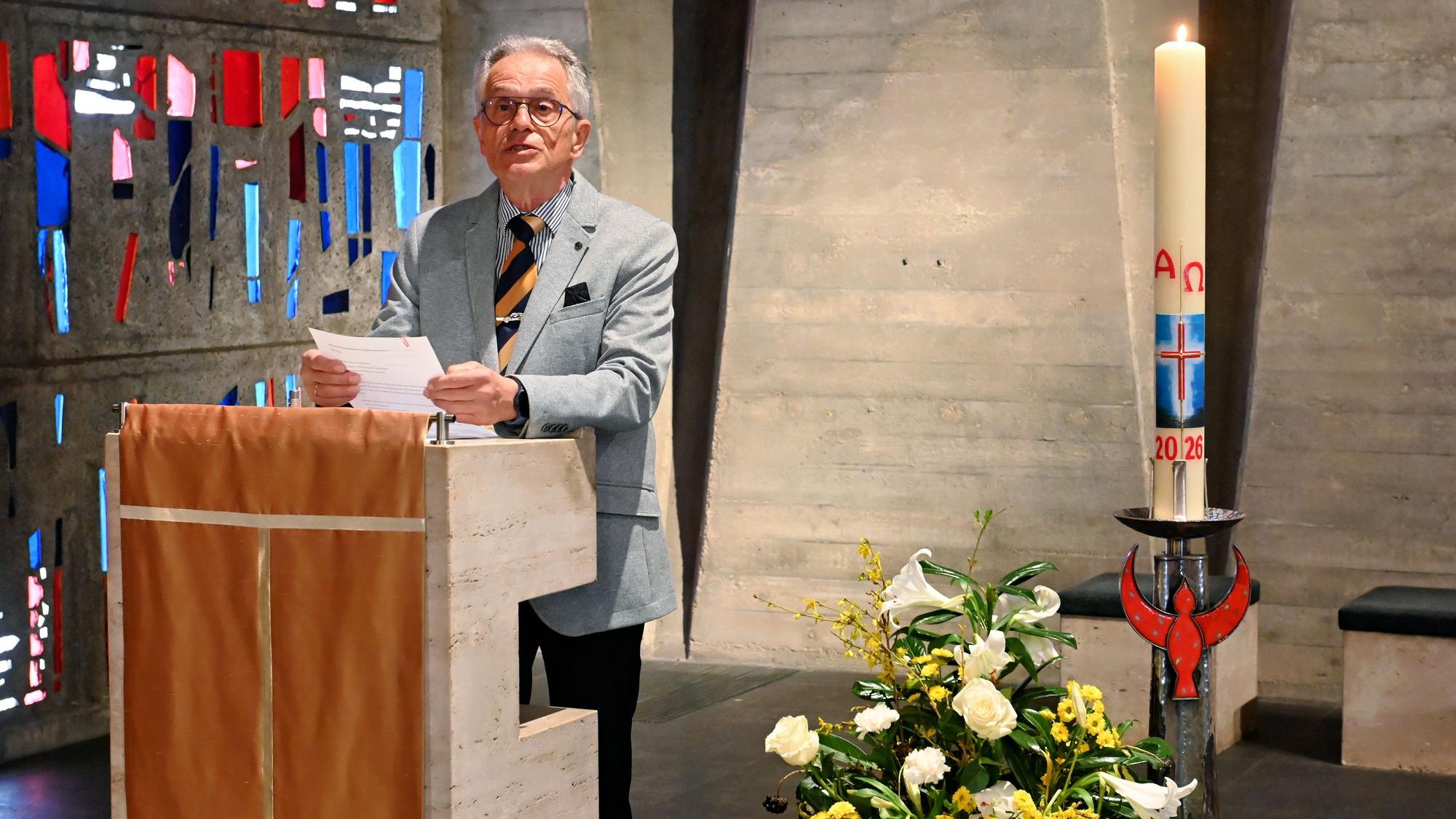 Redner Dr. Hans-Josef Bastian steht in der Kapelle, hinter ihm die bunten Glasfenster, rechts neben ihm ein Blumengesteck und eine brennende Kerze.