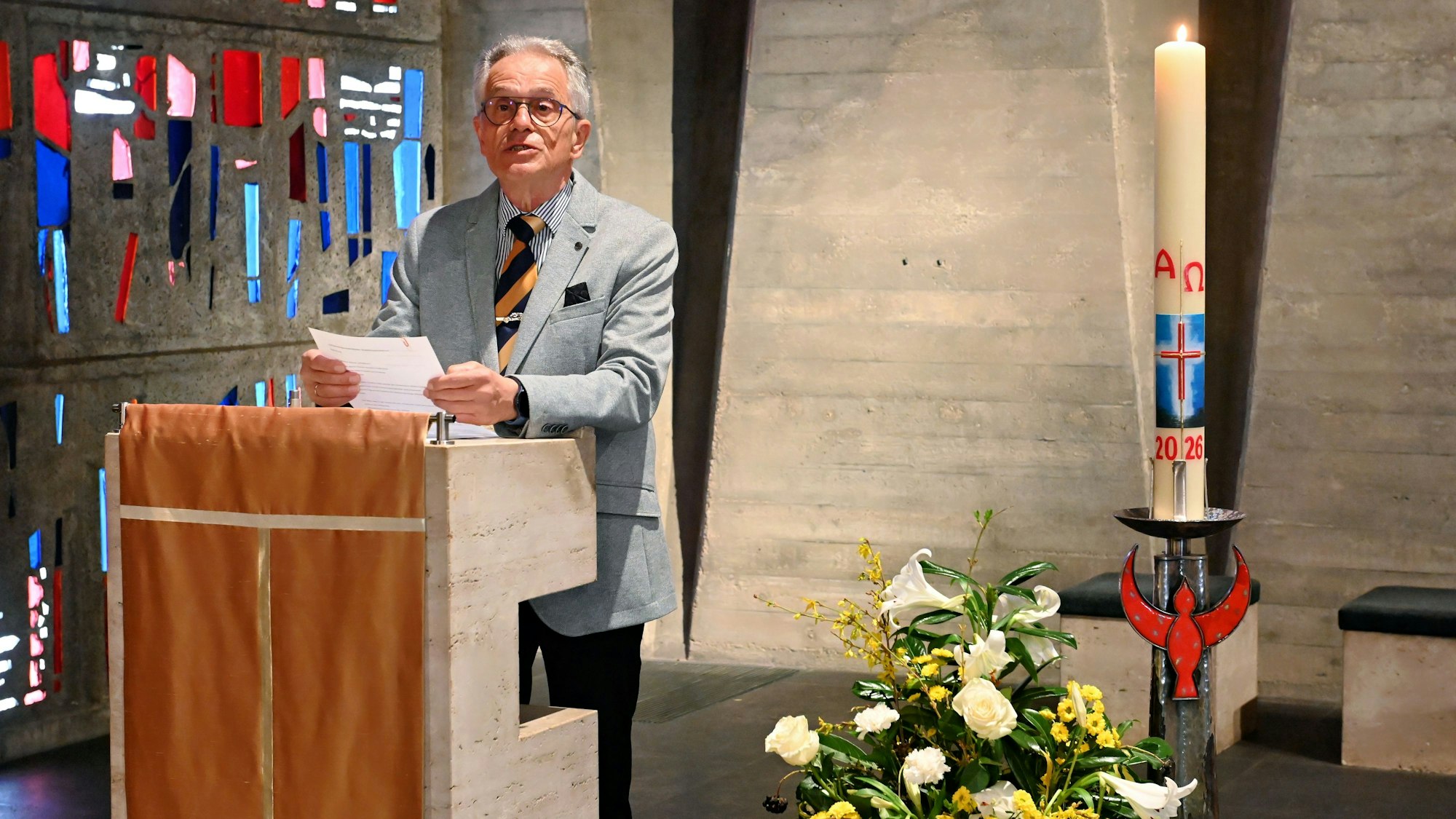 Redner Dr. Hans-Josef Bastian steht in der Kapelle, hinter ihm die bunten Glasfenster, rechts neben ihm ein Blumengesteck und eine brennende Kerze.