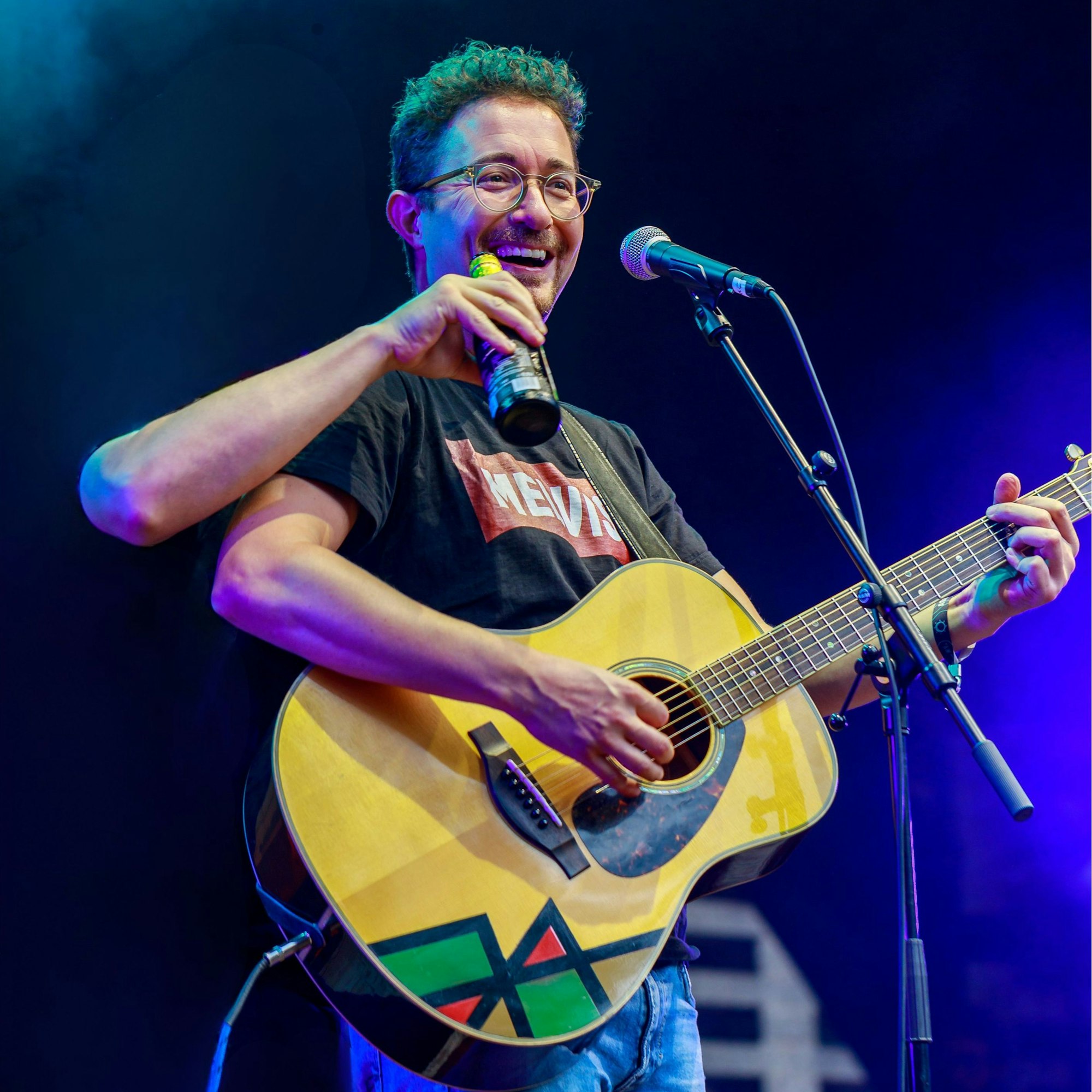 Melvin Haack steht Gitarre spielend auf der Bühne vor einem Mikrophonständer. Aus der Dunkelheit hinter ihm kommt ein dritter Arm und hält ihm eine Bierflasche vor den Mund.