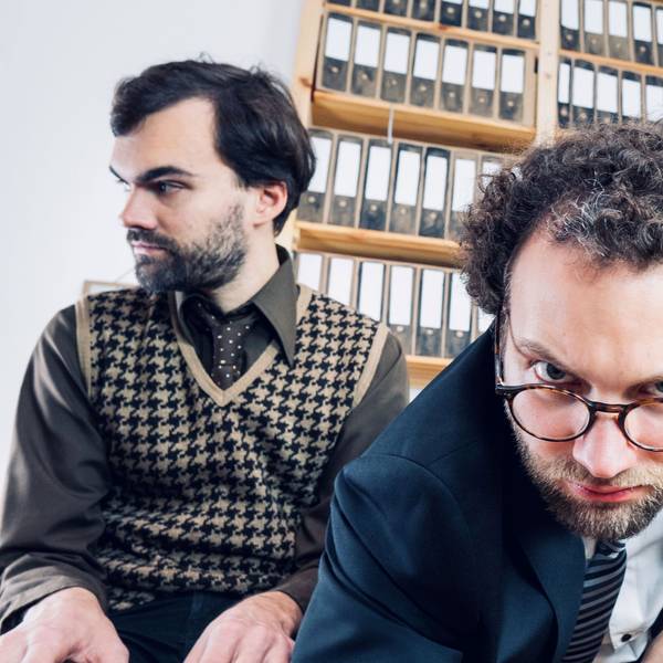 Drei junge Männer in Businesskleidung sitze vor einer Wand voller Aktenordner. Der Mann in der Mitte beugt sich in Richtung Kamera.