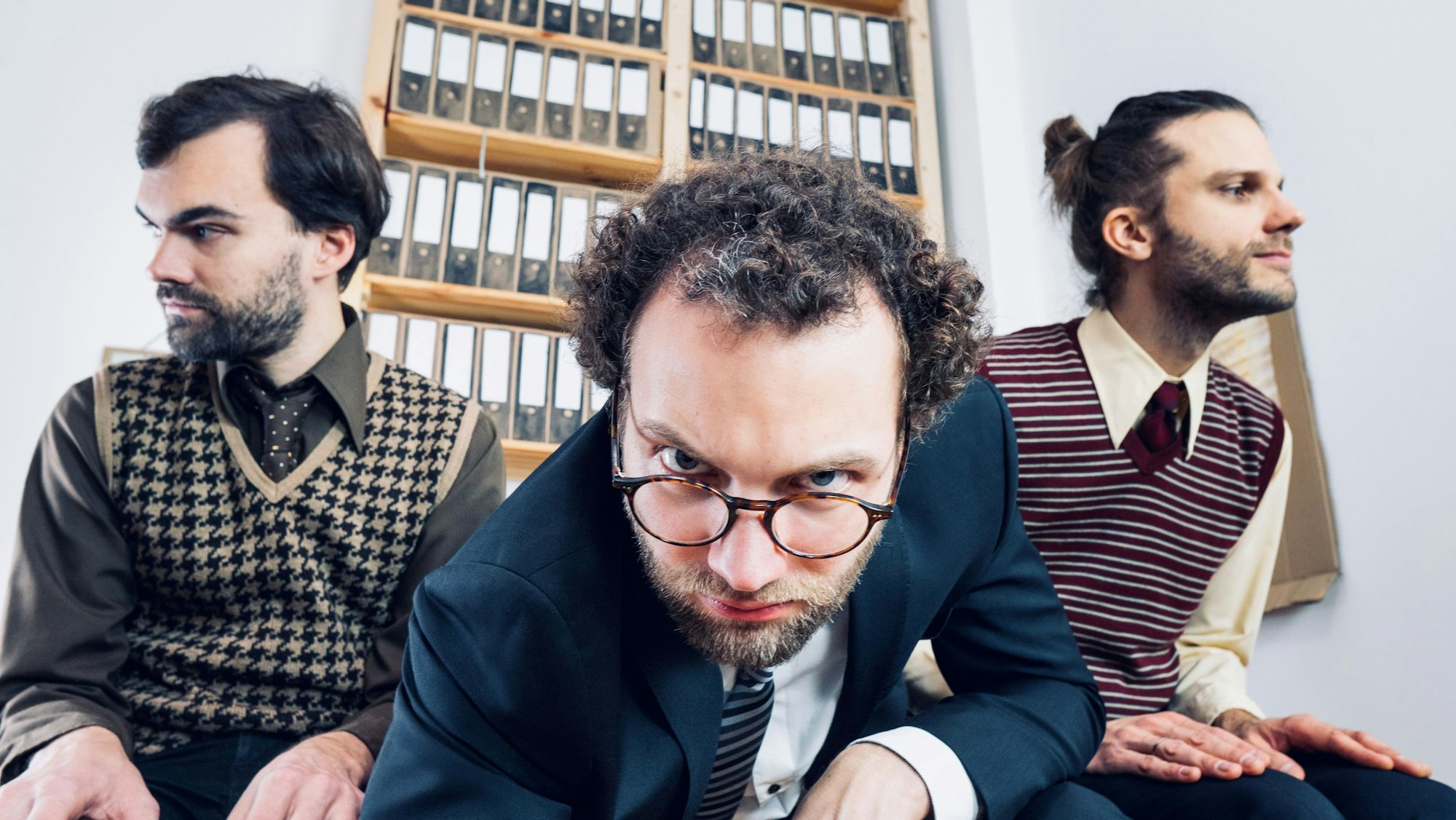 Drei junge Männer in Businesskleidung sitze vor einer Wand voller Aktenordner. Der Mann in der Mitte beugt sich in Richtung Kamera.