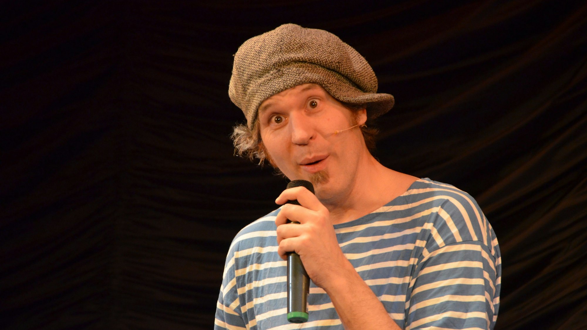 Drop Bert hat ein Streifen-Shirt an und eine Kappe schief auf dem Kopf. Er spricht in sein Mikro, das er in der Hand hält und reißt die Augen weit auf.