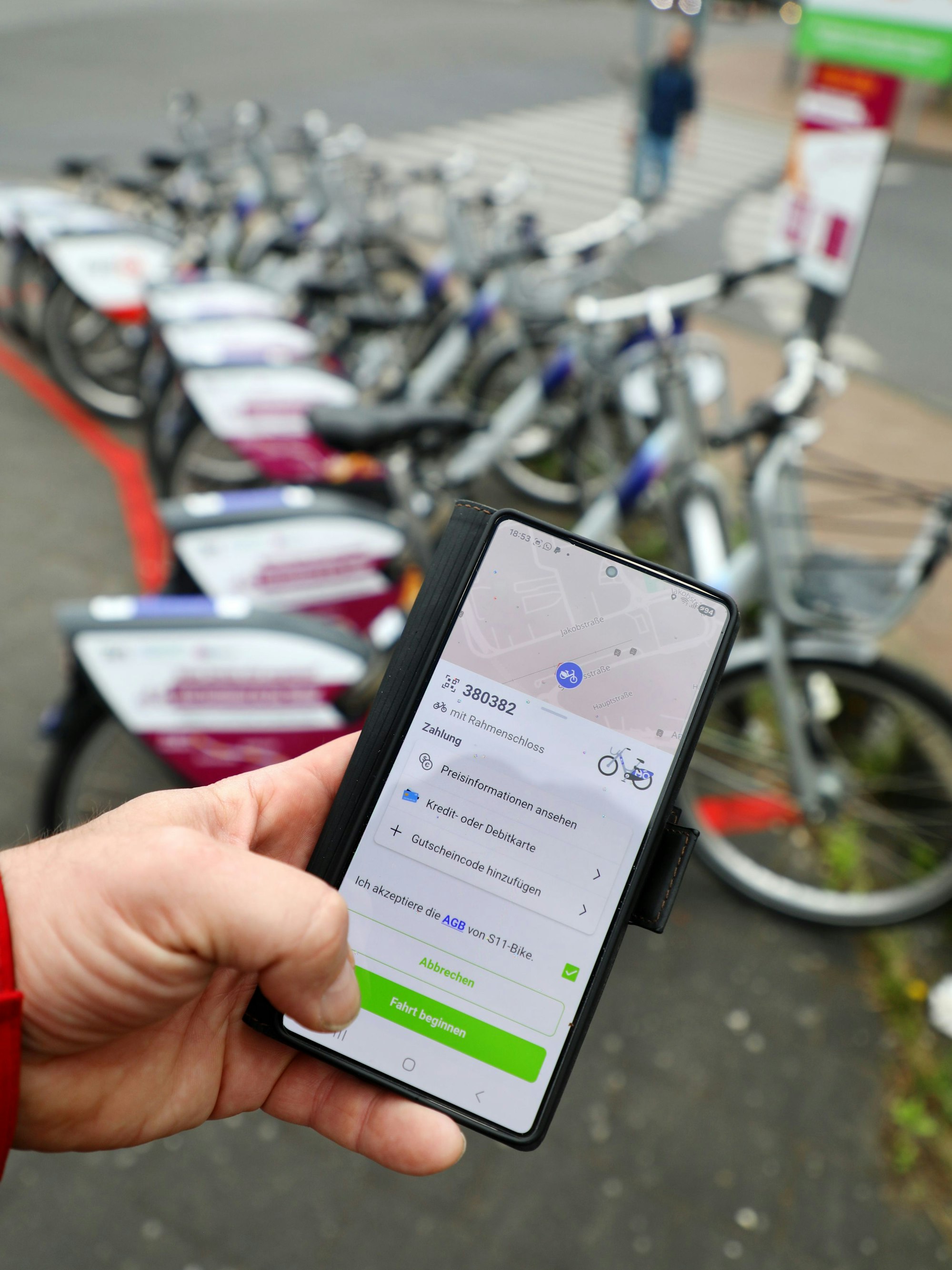 Eine Hand hält ein Handy mit einer App, mittels derer eines von mehreren Fahrrädern im Hintergrund ausgeliehen wird.