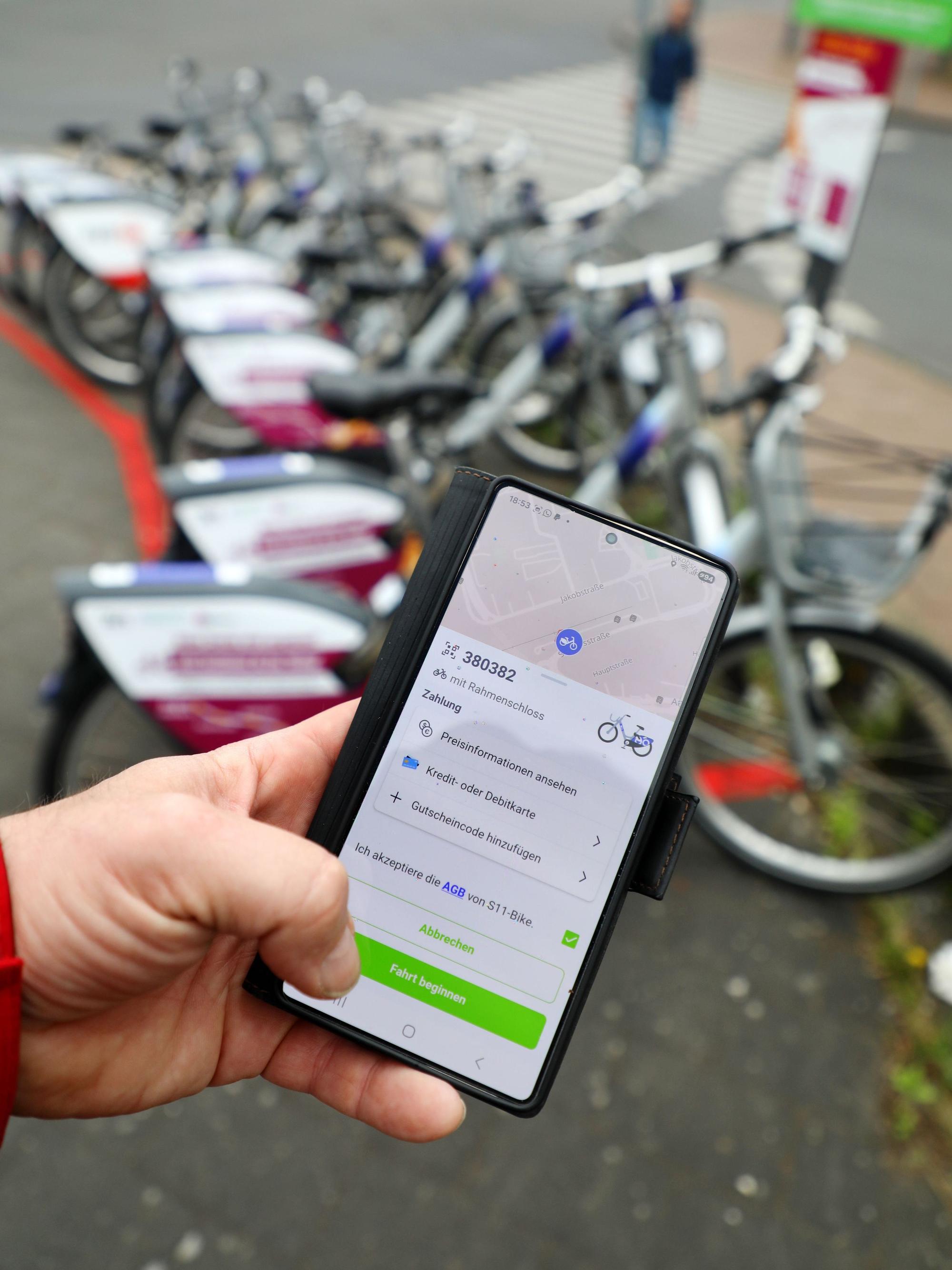 Eine Hand hält ein Handy mit der App zum Ausleihen der SEV-Leifahrräder am Bergisch Gladbacher S-Bahnhof.