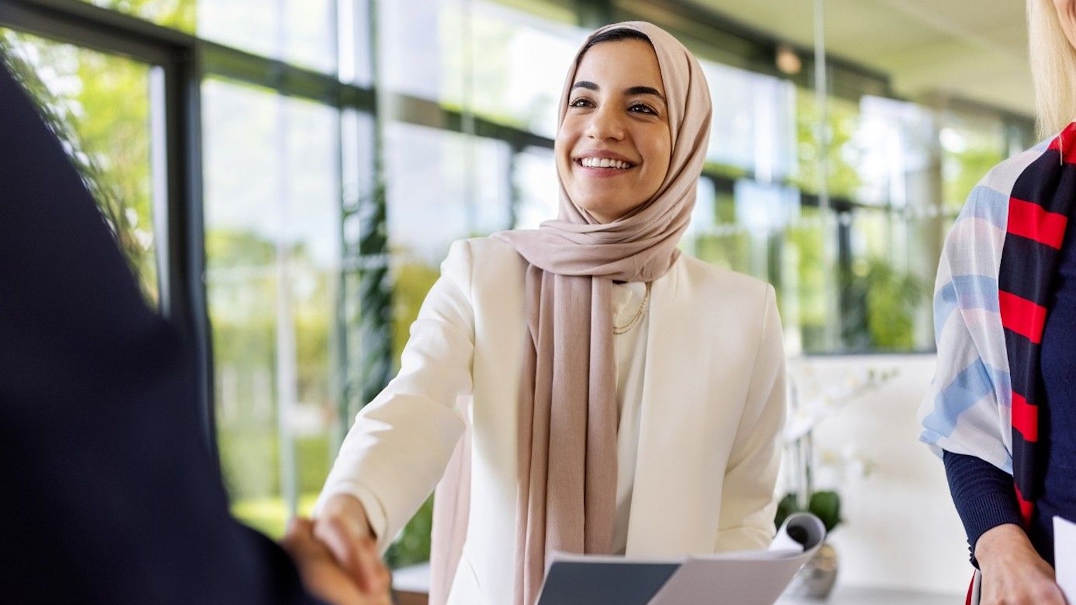 Junge Frau schüttelt einer Person gegenüber die Hand und lächelt. Sie hat Bewerbungsunterlagen in der Hand.