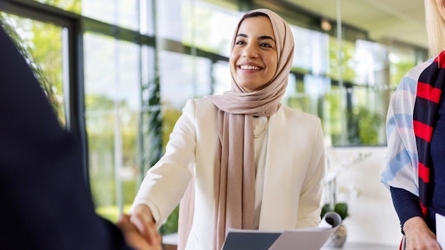 Junge Frau schüttelt einer Person gegenüber die Hand und lächelt. Sie hat Bewerbungsunterlagen in der Hand.