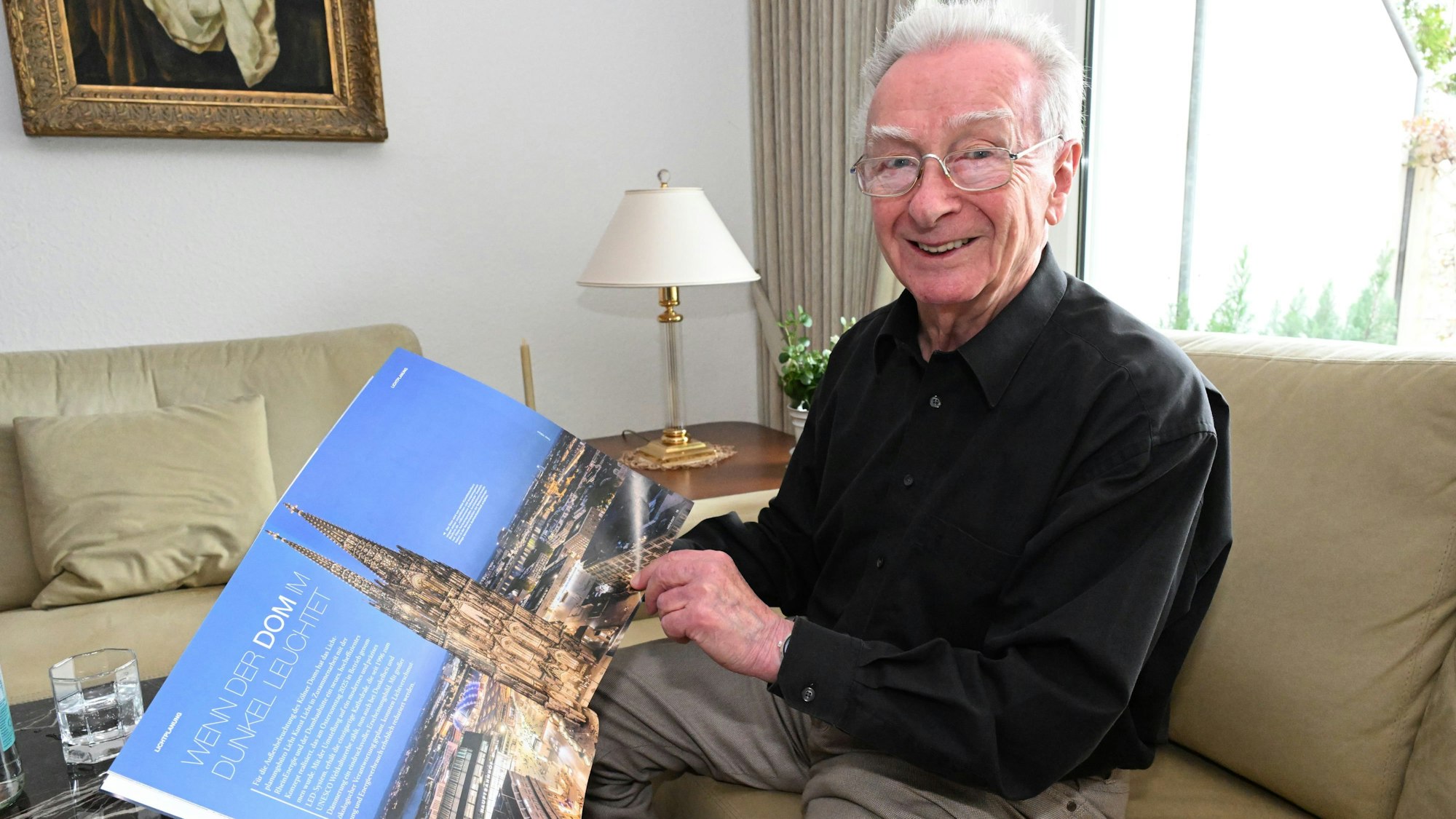 Ein älterer Herr sitzt auf einem Sofa und hält ein Heft mit einem Hochglanzfoto vom beleuchteten Klölner Dom in die Kamera.