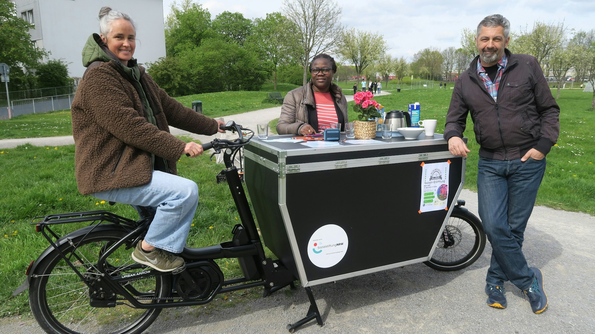 Eine Frau sitzt auf dem Sattel eines stehenden Lastenrads. Auf dem zu einem Tisch umgebauten Lastenteil stehen eine dunkelhäutige Frau und ein Mann.