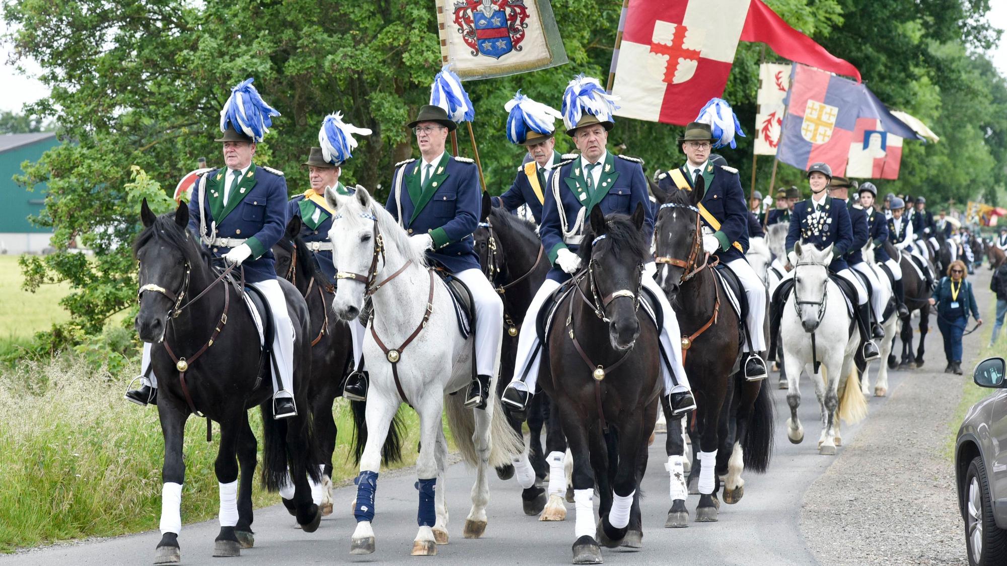 Zahlreiche Männer zu Pferd reiten über einen Feldweg.