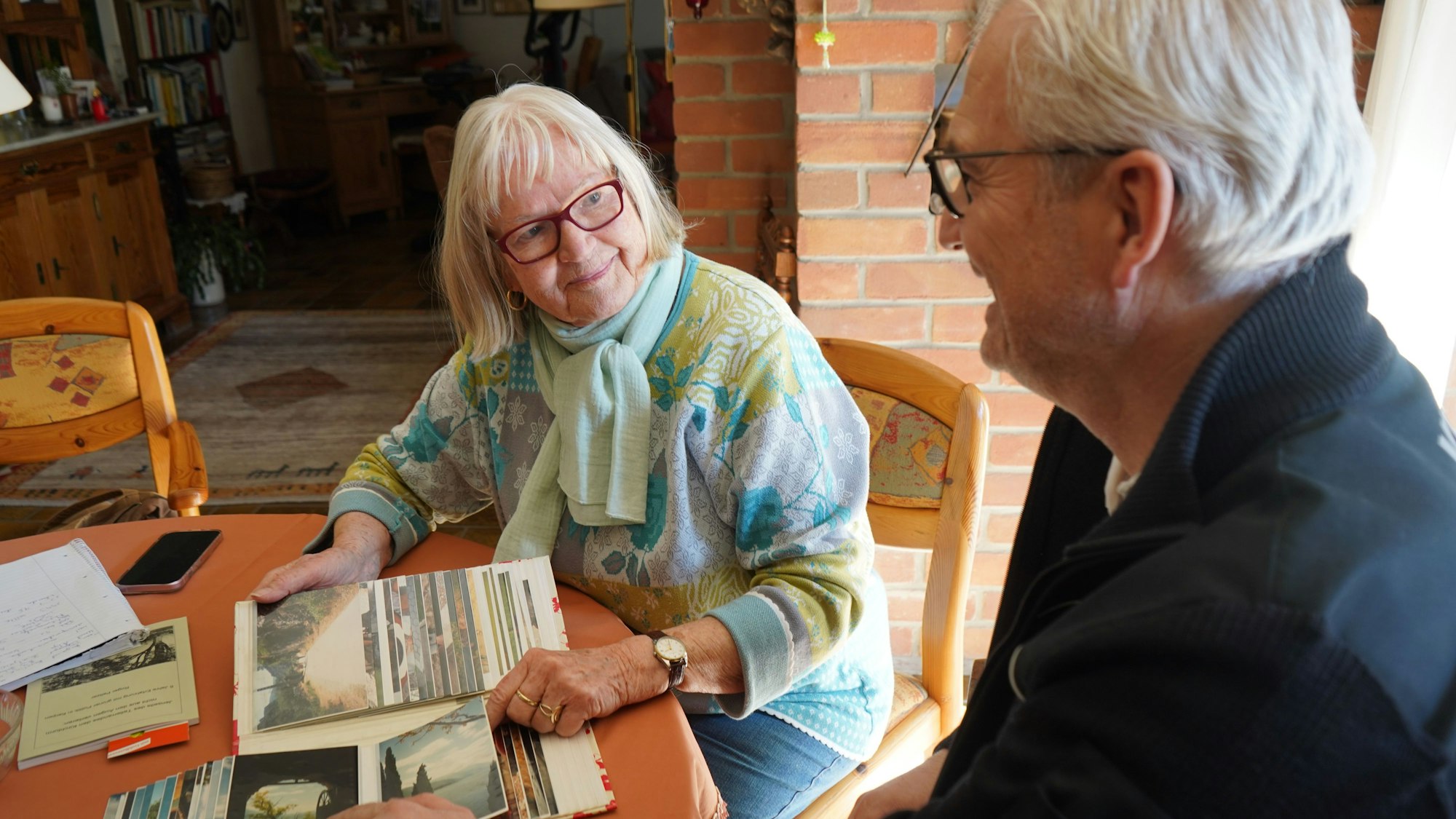 Das Foto zeigt eine ältere Frau und einen Mann, die sich Fotos auf einem Tisch anschauen.
